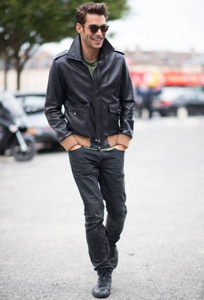 El hombre modelo elegante y guapo de moda con ropa de moda con chaqueta de  cuero y gafas de sol lleva una capucha y camina por la ciudad