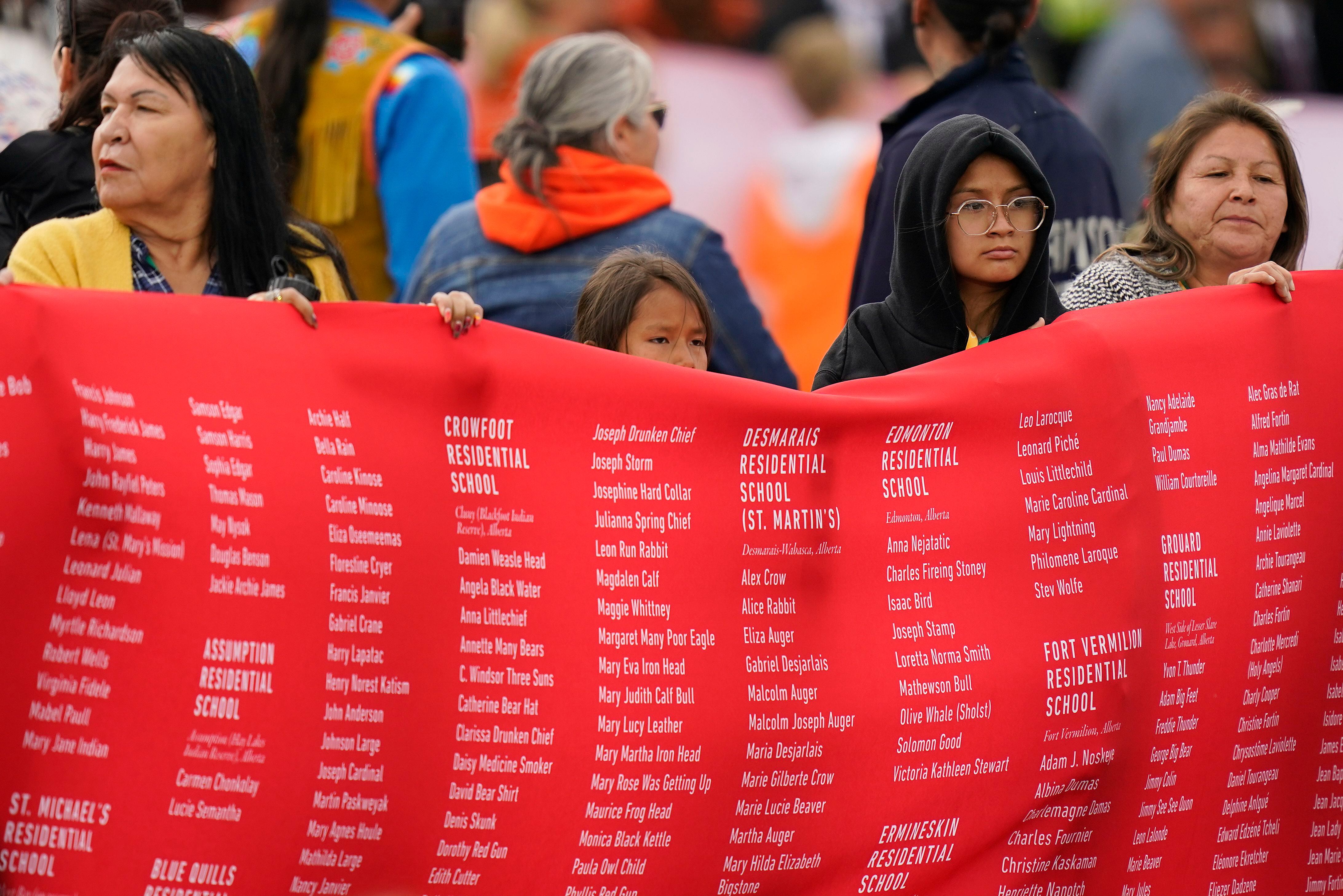 Mujeres indígenas sostienen el telón rojo que lleva inscritos los nombres de los menores fallecidos en la red de internados. Las palizas, las agresiones sexuales, el abandono, el racismo y el repudio cultural fueron algunas de las prácticas denunciadas en esos internados.
