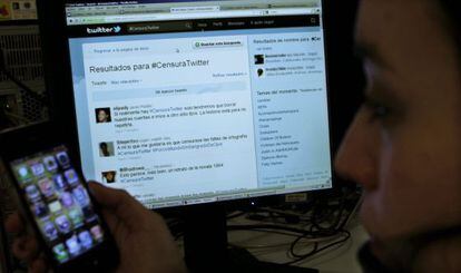 Un usuario de Twitter mira su m&oacute;vil ante un ordenador conectado a un apartado de la red social en el que los tuiteros critican a la empresa.