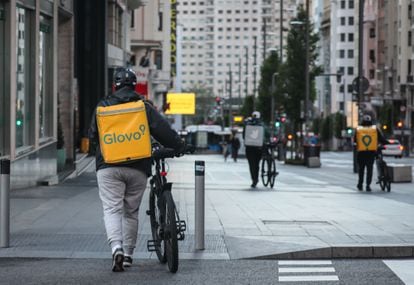 Dos repartidores de Glovo y otro de Deliveroo caminan junto a sus bibicletas por la acera de la Gran Vía de Madrid.