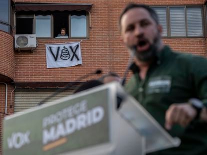 Santiago Abascal en la concentración de Vox en la Plaza de la Constitución en Vallecas, y en el fondo una vecina con una pancarta de protesta.