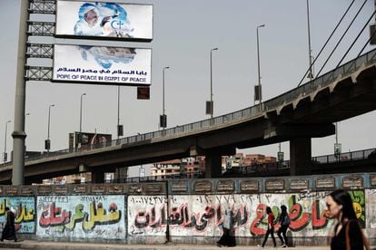 Ciudadanos egipcios pasan por delante de una pared pintada con un grafiti sobre la cual se puede ver un cartel que anuncia la visita del papa Francisco a El Cairo (Egipto).
