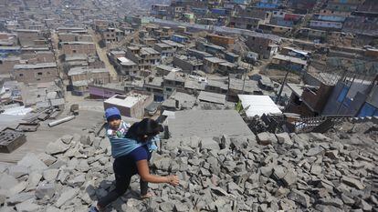 Una mujer lleva a su hijo a la espalda y camina sobre las rocas en chanclas hacia una casa donde se distribuyen alimentos, en Lima (Perú).