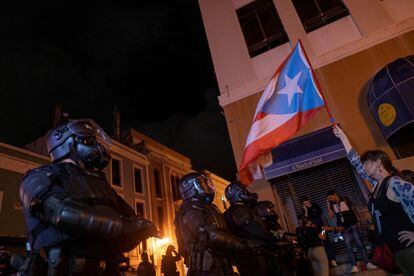 En un comunicado, la Policía de Puerto Rico informó este viernes que cinco agentes resultaron heridos y cuatro personas fueron detenidas durante las protestas. En la imagen, una manifestante sostiene una bandera puertorriqueña frente a un grupo de policías antidisturbios.