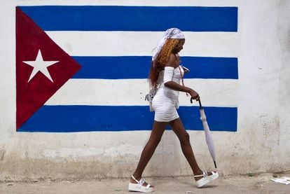 Una mujer vestida de blanco (practicante de la religión yoruba) camina frente a un grafiti de la bandera cubana.