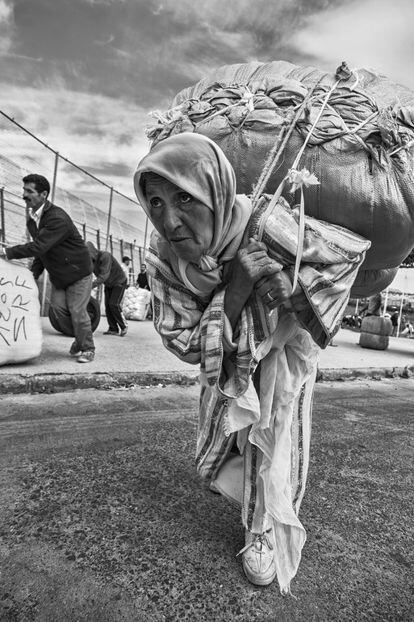 Una mujer porteadora se dirige hacia la frontera entre Marruecos y España con un pesado bulto a la espalda. Tras ella, algunos hombres hacen el mismo trayecto. Los pocos hombres que participan en este comercio irregular de mercancías no cargan bultos sobre la espalda. Se limitan a empujarlos y hacerlos rodar. La mayoría de mujeres tendrá que caminar hasta las naves del lejano polígono, cargar sobre sus lumbares y alcanzar la valla.