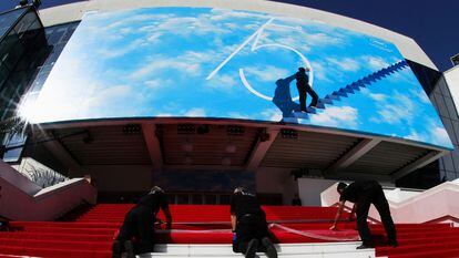 Tres operarios instalan la alfombra roja del Festival de Cannes a la entrada del Palacio de Festivales, decorado con el póster del certamen, que homenajea a 'El show de Truman'.