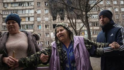 People help an elderly woman to walk in a street with an apartment building hit by shelling in the background in Mariupol, Ukraine, Monday, March 7, 2022. (AP Photo/Evgeniy Maloletka)