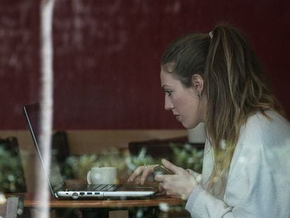 Una joven consulta su dispositivo móvil y su portátil a la vez mientras desayuna en el interior de una cafetería.