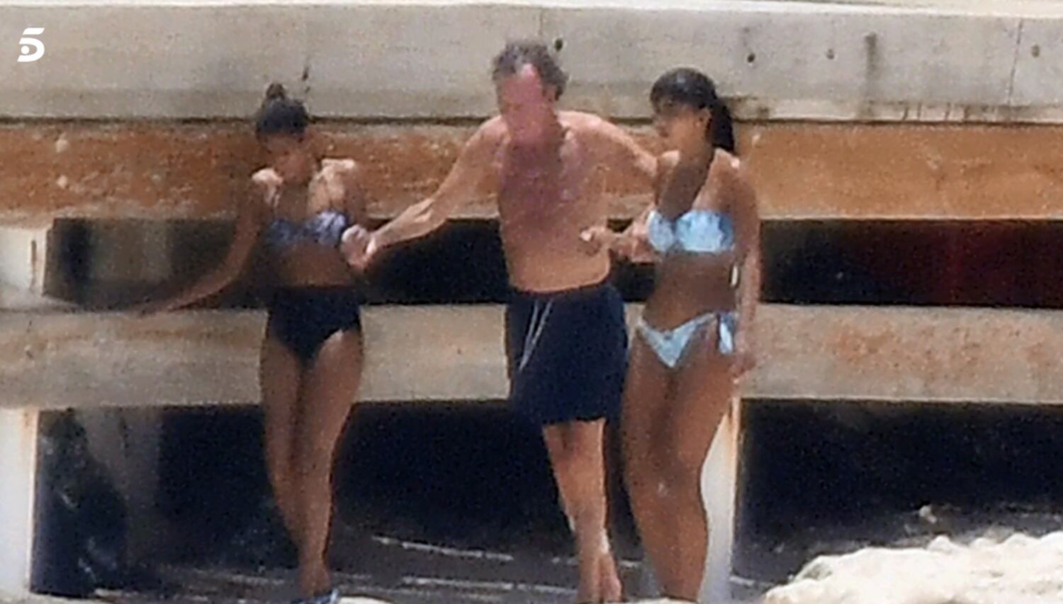 Julio iglesias caminando por la playa ayudado por dos mujeres.