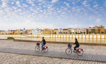 Dos turistas en bicis públicas de alquiler pedaleando junto al río Guadalquivir, en Sevilla, con el barrio de Triana al fondo.