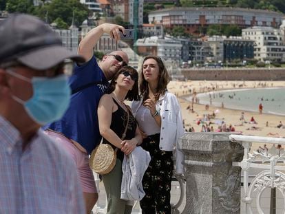 Un señor con mascarilla pasa por delante de un grupo de turistas que se fotografían en la playa donostiarra de La Concha.