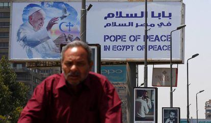Un hombre conduce su bicicleta en el cartel de la visita del Papa a El Cairo al fondo. 