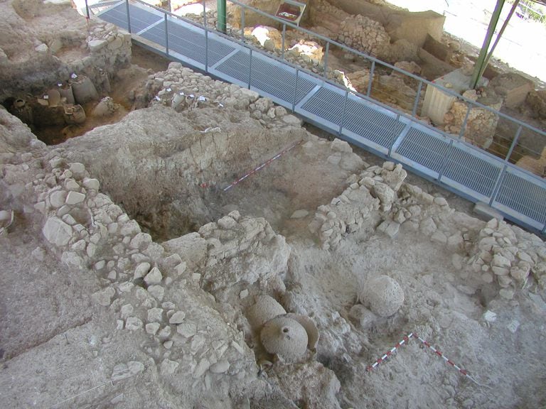 Vivienda íbera excavada con una pasarela que cruza una calle de la población. En primer plano, dos molinos rotatorios de piedra.