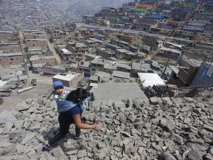 Una mujer lleva a su hijo a la espalda y camina sobre las rocas en chanclas hacia una casa donde se distribuyen alimentos, en Lima (Perú).