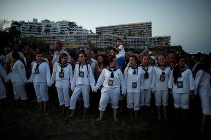 Mujeres vestidas con trajes tradicionales contemplan la subida en barca de la Virgen del Carmen desde la playa.