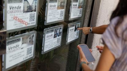 Dos personas observan varios anuncios de venta de viviendas en un escaparate de una inmobiliaria, en Madrid.
