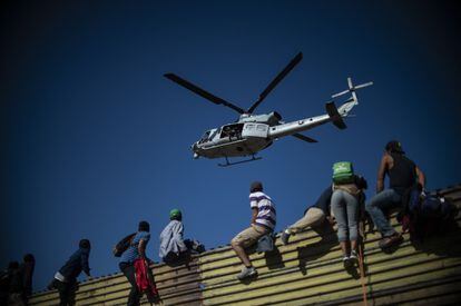 Varias personas en lo alto de la valla fronteriza observan un helicóptero de la policía de los EE.UU.