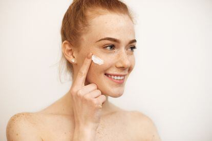 Lovely caucasian girl with red hair and freckles is smiling while using a cream on her face