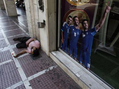 Un hombre pide dinero en una calle de Atenas frente a un cartel que promociona a la selecci&oacute;n de Grecia.