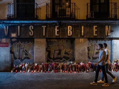 Dos personas pasan frente a las flores en homenaje a Conchi, asesinada en su tienda de la plaza de Tirso de Molina.