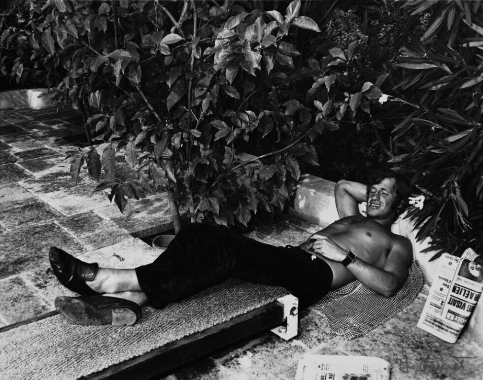 Jean Paul Belmondo descansando sobre un banco con cobertura de rafia en la piscina del hotel. |
