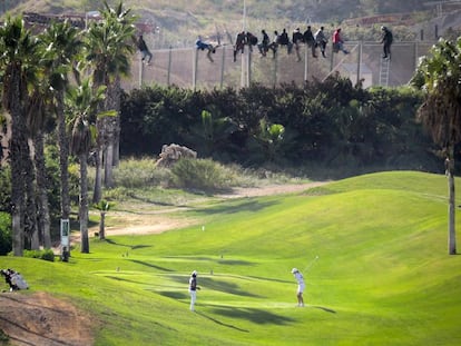 Una docena de inmigrantes permanece encaramado a la valla de Melilla mientras dos personas juegan al golf.