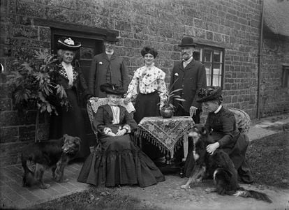 Portrait of a Victorian-era family from Hellidon, Northamptonshire, United Kingdom, posing at the door of their house next to a table with a tablecloth and an aspidistra. 