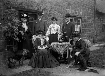 Portrait of a Victorian-era family from Hellidon, Northamptonshire, United Kingdom, posing at the door of their house next to a table with a tablecloth and an aspidistra. 