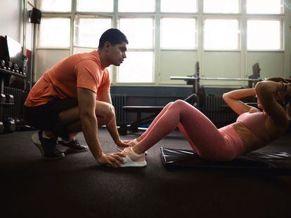 Una joven hace abdominales en un gimnasio, ayudada de su entrenador.