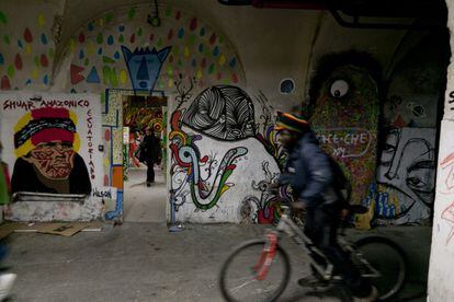 Un hombre se mueve en bicicleta por un pasillo de La Tabacalera, con las paredes decoradas con pinturas y ante una habitación donde se ve a una mujer. El centro cuenta con un taller de arreglo de bicicletas y es común ver este tipo de vehículos apartados en su interior.