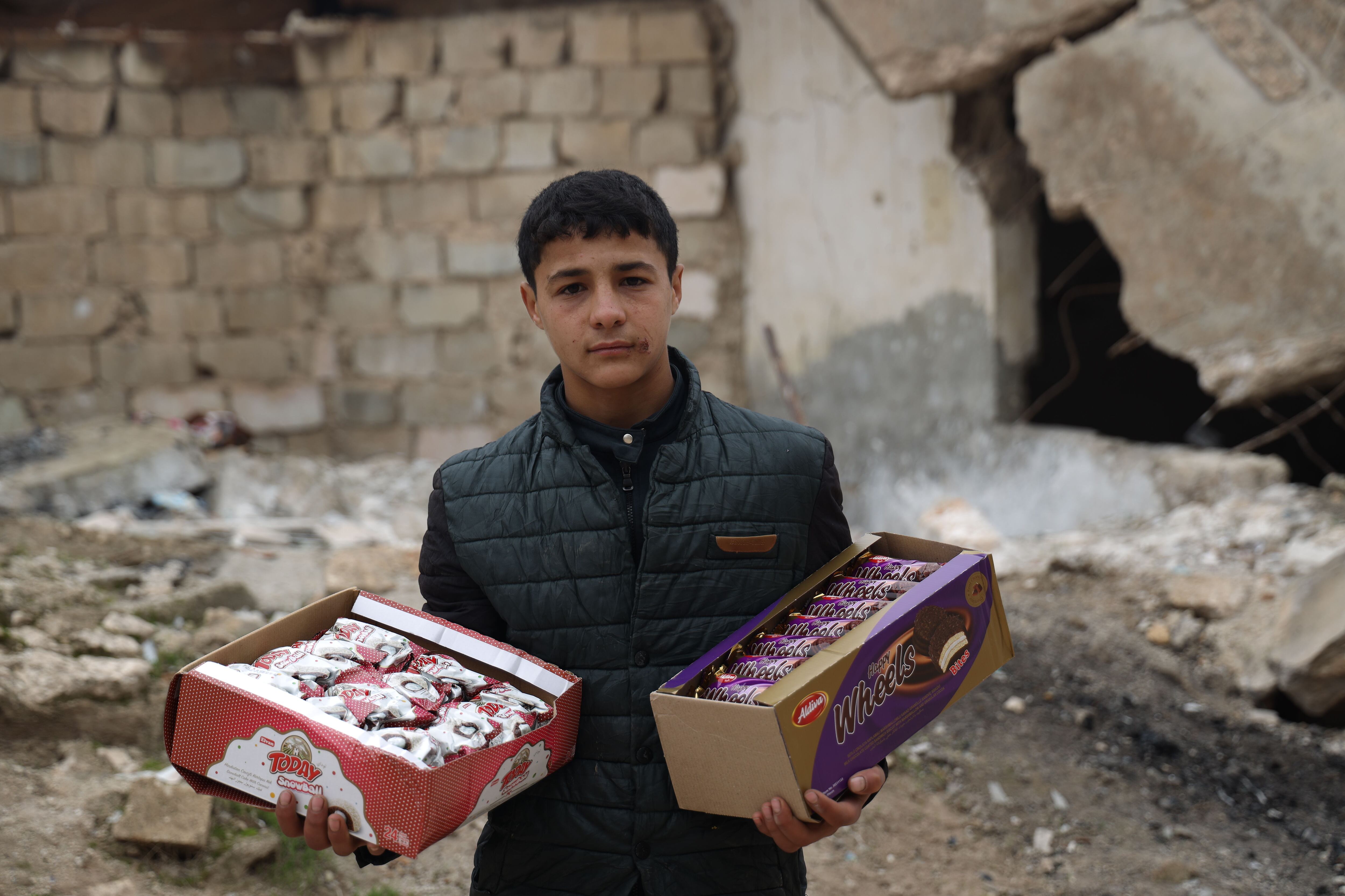 Hussein Mutawa, de 12 años, frente a un edificio destruido en su pueblo de Sarmin, en el campo de Idlib, mientras deambula por las inseguras calles de la ciudad para vender galletas. Hussein dejó la escuela temprano y comenzó este trabajo para ayudar a su madre y sus cinco hermanas.