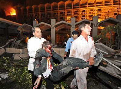 Dos paquistaníes trasladan a una de las víctimas del atentado. Al fondo, la fachada en llamas del hotel Marriott, en el centro de Islamabad.