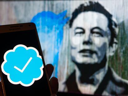 Un móvil muestra el controvertido símbolo de verificación de pago de Twitter, que Elon Musk (al fondo en una pintada en Bruselas, Bélgica) implantó a su llegada a la plataforma.