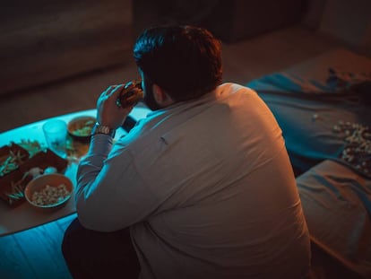 Un hombre comiendo una hamburguesa delante de la televisión en una imagen de stock.