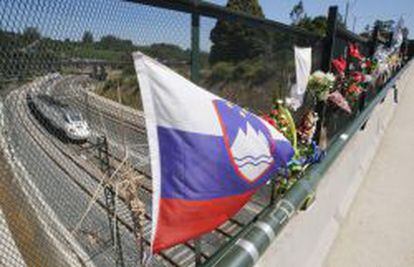 Ramos de flores y una bandera de Eslovenia recuerdan a las v&iacute;ctimas del accidente ferroviario del 24 de julio del a&ntilde;o pasado. De fondo, un tren de velocidad alta circula por la curva de A Grandeira.