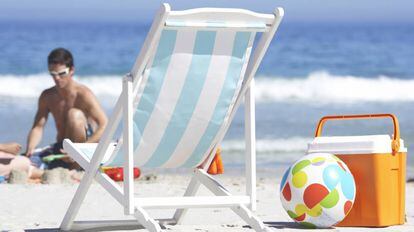 Una nevera de playa y un balón inflable descansan al lado de una silla en la orilla.