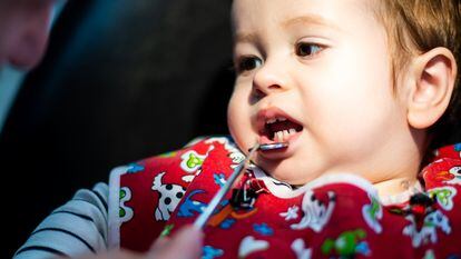 Un niño de menos de dos años en la consulta del dentista, con una bata roja, mira al dentista mientras este le baja el labio inferior con un pequeño espejo.
