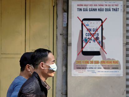Dos hombres protegidos con mascarillas pasan frente a un cartel en Hanoi (Vietnam) que advierte contra la difusión de bulos.