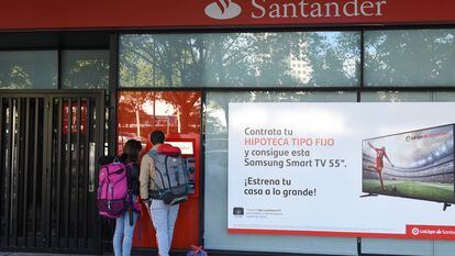 Dos personas retiran dinero de un cajero automático junto a un anuncio de hipotecas del Banco Santander, en una oficina de Madrid.