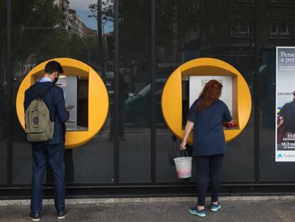 Una señora limpia un cajero de CaixaBank mientras un chico realiza una operación en la calle Orense (Madrid).