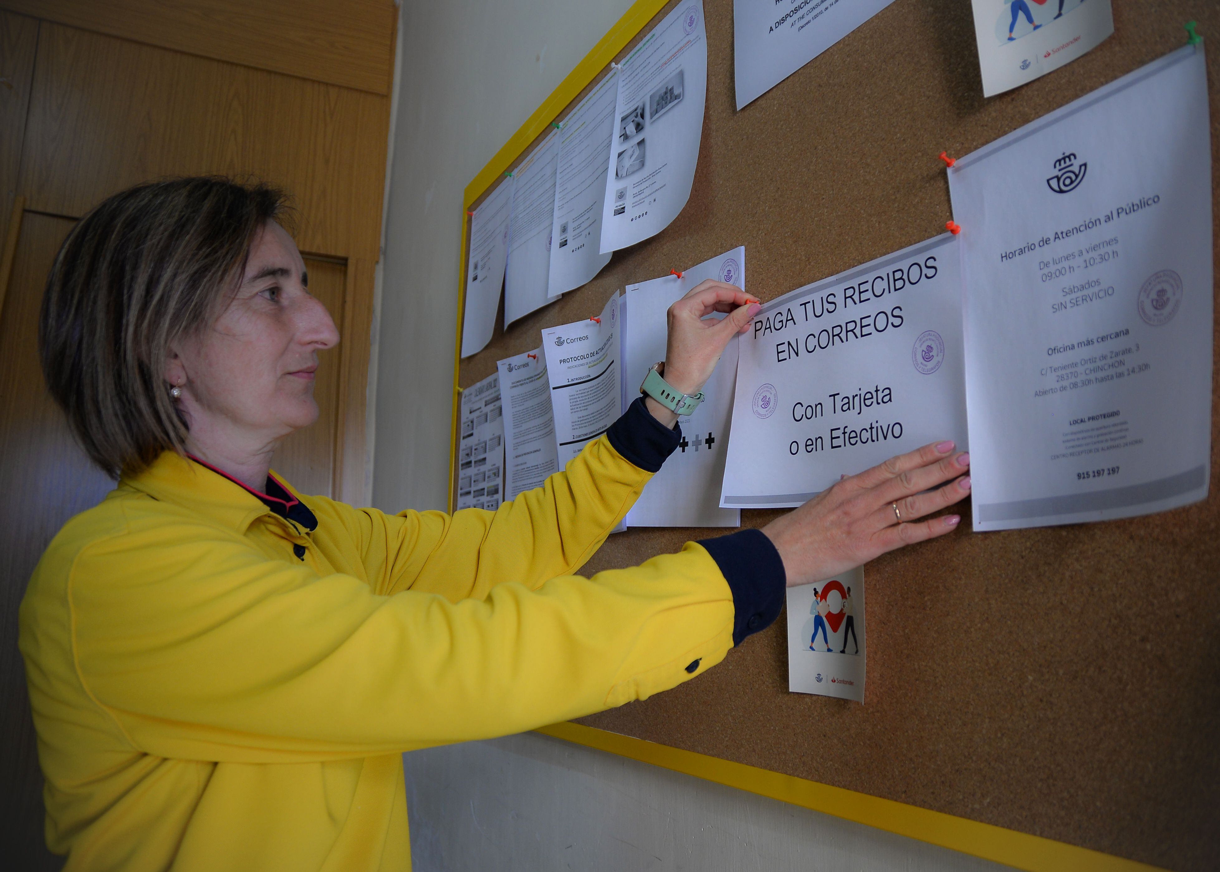 La cartera rural María del Carmen Ávila cuelga en el tablón de la oficina de Correos de Belmonte de Tajo un cartel sobre el nuevo servicio de cobro de recibos.