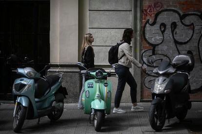 Two young men walk along a narrow sidewalk in Barcelona where several motorcycles are parked. 