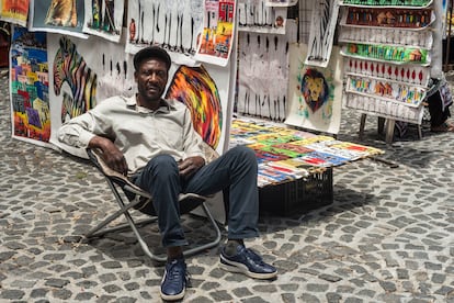 Peter, artista congoleño residente en Sudáfrica desde hace más de dos décadas, posa en su silla de camping, junto a sus pinturas, en la plaza de Green Market, en Ciudad del Cabo. Pincha en la imagen para ver la fotogalería completa.