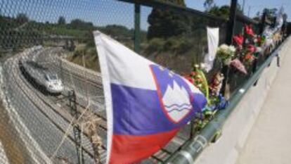 Ramos de flores y una bandera de Eslovenia recuerdan a las v&iacute;ctimas del accidente ferroviario del 24 de julio del a&ntilde;o pasado. De fondo, un tren de velocidad alta circula por la curva de A Grandeira.