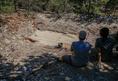 Dos estudiantes descansan frente a la Cata Botánicos mientras el dron sobrevuela la superficie descubierta para documentarla. La sombra del aparato se aprecia en la izquierda superior del perímetro de estudio.