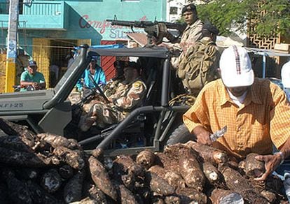 Un hombre vende yuca en un mercado de Santo Domingo mientras una patrulla militar vigila la zona.