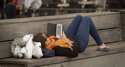 Una joven lee un libro electr&oacute;nico tumbada en un banco, en Sevilla. 
