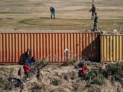 Un pequeño grupo de migrantes se topa con un muro de contenedores a su llegada a Eagle Pass, en Texas. Del lado estadounidense, un grupo de hombres juegan golf.