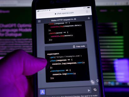 Una persona maneja un teléfono móvil con un programa de inteligencia artificial. Al fondo, una pantalla de ordenador con la página web de ChatGPT.